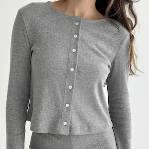 Könnte beinhalten: Graue Strickjacke mit Rippenmuster, langen Ärmeln und einer Reihe weißer Knöpfe auf der Vorderseite. Die Strickjacke ist Teil eines passenden Sets. Der Stoff hat vertikale Linien und der Ausschnitt ist ein Rundhalsausschnitt.