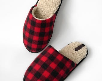 May include: A pair of slip-on slippers featuring a red and black buffalo plaid pattern on the exterior. The interior is lined with a cream-colored, textured fleece material. Each slipper has a small dark tag near the heel area.