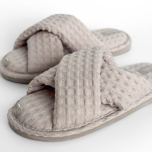 May include: A pair of light taupe cross-strap slippers. The slippers have a textured waffle weave pattern and a thick sole. The design features a crisscross strap across the top of the foot. The background is white.