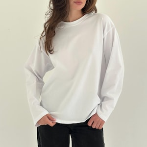 Pode incluir: Uma camisa branca de manga comprida com gola redonda. A camisa é feita de um tecido macio e opaco. As mangas são compridas e folgadas. A camisa é combinada com calças pretas.