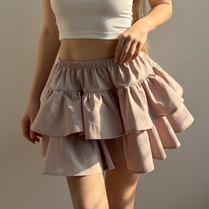 Könnte beinhalten: Ein hellbrauner, gestufter Rüschenrock. Der Rock hat einen elastischen Bund und ein mehrschichtiges Design. Der Stoff scheint ein weiches, fließendes Material zu sein. Der Rock ist eine neutrale Farbe, passend für verschiedene Outfits.