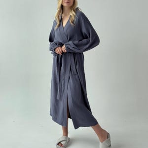 May include: A long, slate blue robe with a wrap-around design and a matching belt. The robe features long sleeves with gathered cuffs and a V-neckline. The fabric appears to be a lightweight, textured material. The model is wearing gray slippers.