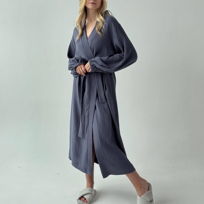 May include: A long, slate blue robe with a wrap-around design and a matching belt. The robe features long sleeves with gathered cuffs and a V-neckline. The fabric appears to be a lightweight, textured material. The model is wearing gray slippers.