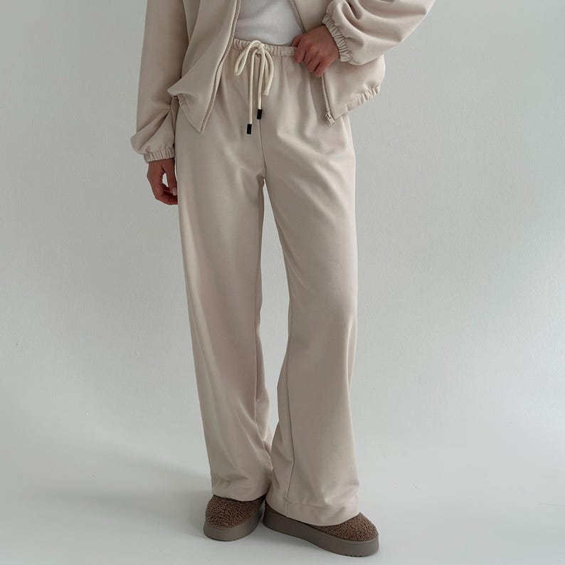 May include: A beige two-piece tracksuit. The jacket has a zipper and the pants have a drawstring. The pants are wide-legged. The shoes are brown and fuzzy.