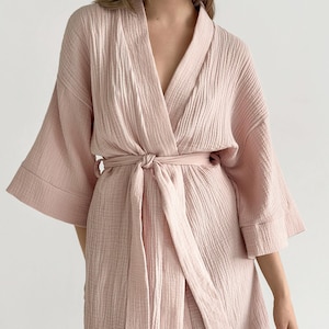 May include: A light pink robe made of textured muslin fabric featuring wide, three-quarter length sleeves, a V-neckline, and a matching fabric tie belt at the waist. The garment has a relaxed fit with visible vertical crinkle patterns throughout.