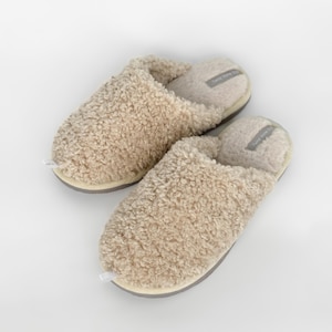 May include: A pair of beige, fluffy slippers with a textured, curly exterior. The slippers have a light grey sole and a soft, cushioned interior. The tag on the inside of the slipper reads "Twice Home Co."