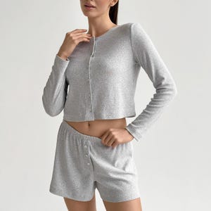 Könnte beinhalten: Ein hellgraues Pyjama-Set aus Rippstrick. Das Set besteht aus einer langärmligen, geknöpften Strickjacke und passenden Shorts mit Knopfverschluss. Der Stoff wirkt weich und bequem, geeignet für Nachtwäsche oder Loungewear.