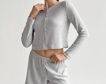 May include: A light gray, ribbed knit pajama set. The set includes a long-sleeved, button-down cardigan and matching shorts with a button closure. The fabric appears soft and comfortable, suitable for sleepwear or loungewear.