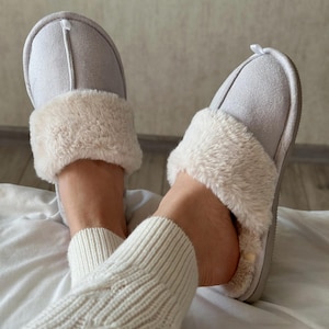 May include: A pair of light gray suede slippers with a fluffy, cream-colored faux fur trim. The slippers have a soft, cushioned sole and are designed for indoor wear. A white ribbon detail is visible on the back of each slipper.