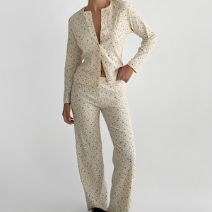 Peut inclure: Un haut à manches longues boutonné et un pantalon droit assorti en tissu crème avec un motif floral à petite échelle dans des tons de bleu, rose et jaune. L'ensemble présente un imprimé cohérent sur tout le vêtement.
