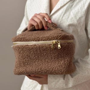 Puede incluir: Un neceser rectangular marrón y esponjoso con cremallera y asa doradas. La bolsa está hecha de un material suave y texturizado, y la sostiene una persona.