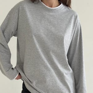 May include: A light grey long-sleeved shirt with a crew neck and a white undershirt collar. The shirt has a relaxed fit and is made of a soft, comfortable material. The sleeves are long and cover the arms.