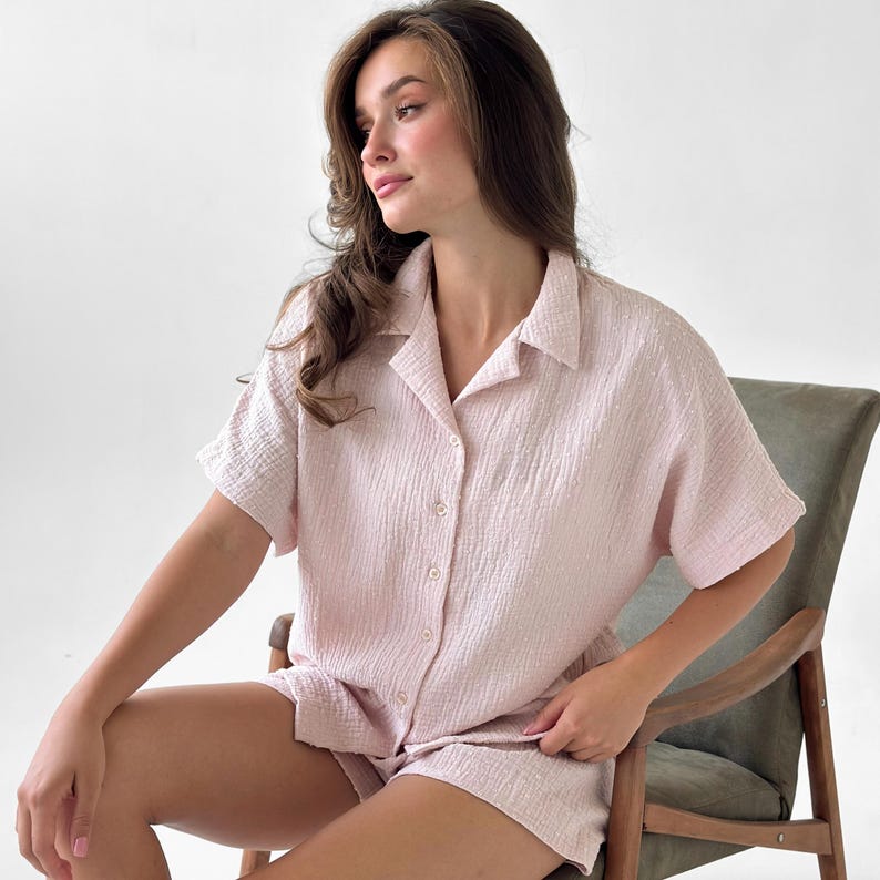 May include: A light pink pajama set featuring a short-sleeved button-down shirt and matching shorts. The set is made of a textured fabric and is designed for comfort and relaxation. The model is sitting in a chair.