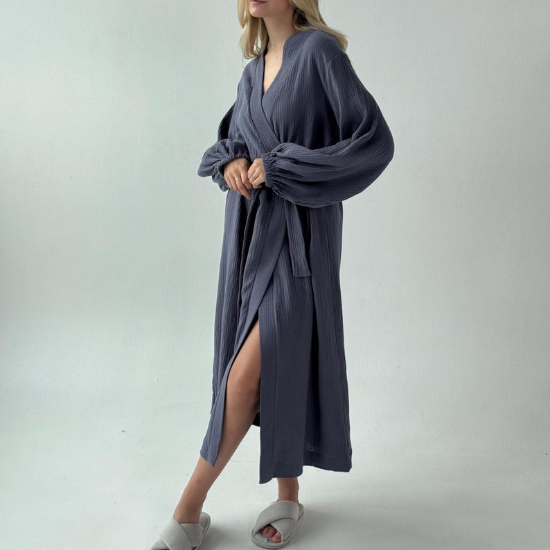 May include: A long, slate blue robe with a wrap-around design and a matching belt. The robe features long, voluminous sleeves and a high side slit. The fabric has a textured appearance. The model is wearing gray slippers.