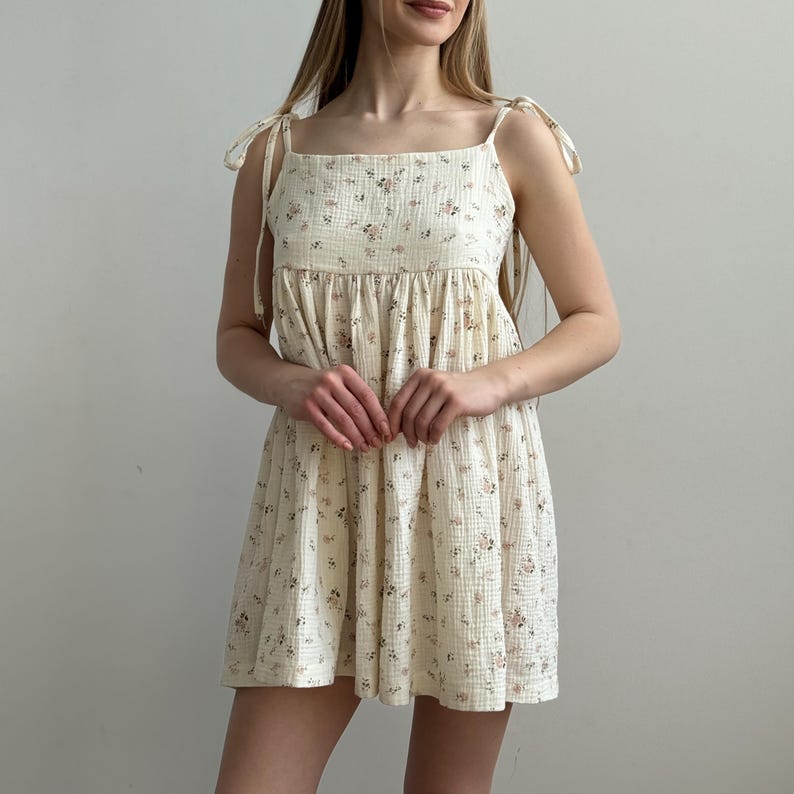 Op de afbeelding: Een cr&egrave;mekleurige, mouwloze jurk met een bloemenpatroon. De jurk heeft strikbandjes en een gelaagde rok. De stof lijkt een lichtgewicht, getextureerd materiaal te zijn, geschikt voor warm weer. De jurk is knielang.