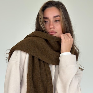 May include: A dark olive green scarf draped over a cream-coloured jacket. The scarf is thick and textured, providing warmth. The jacket has a zip closure and a simple design. The overall look is casual and comfortable, suitable for cooler weather.