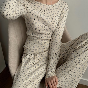 May include: Cream-colored long-sleeved top and matching pants set. The ribbed fabric is covered in a delicate pattern of small, dark blue floral designs. The top has a crew neck, and the pants have a drawstring waist.
