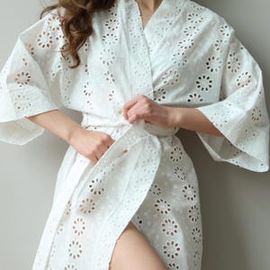 Może przedstawiać: Białe kimono z haftem angielskim i kwiatowym wzorem. Szlafrok ma szerokie rękawy i wiązany pas. Tkanina jest lekka i półprzezroczysta. Stylowy i wygodny dodatek do każdej garderoby.