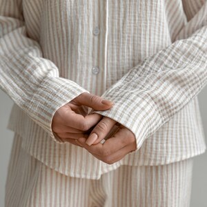 Op de afbeelding: Beige en wit gestreepte pyjamaset. De blouse met lange mouwen heeft een knoopsluiting aan de voorkant en de bijpassende shorts zijn onderaan zichtbaar. De stof heeft een getextureerde, gekreukelde uitstraling. De mouwen zijn bij de polsen gecuffd.