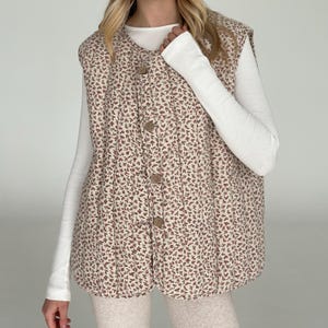 May include: A quilted vest with a floral pattern in shades of beige and burgundy. The vest has a button-up front and a V-neck design. The vest is worn over a white long-sleeved shirt.