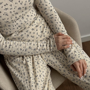 May include: Cream-colored long-sleeved top and pants set with a small, dark floral pattern. The ribbed fabric is visible, and the outfit is worn while seated. The sleeves are long, and the pants have a drawstring waist.