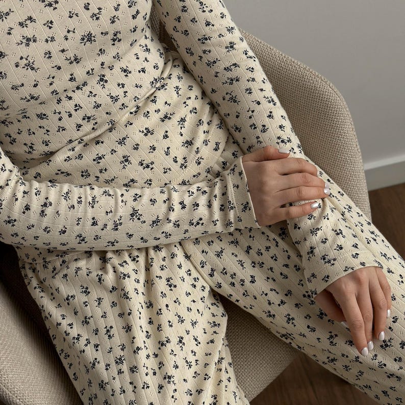 May include: Cream-colored long-sleeved top and pants set with a small, dark floral pattern. The ribbed fabric is visible, and the outfit is worn while seated. The sleeves are long, and the pants have a drawstring waist.