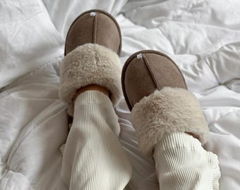 Può includere: Pantofole in camoscio marrone con finiture in ecopelliccia color crema. Le pantofole hanno la punta arrotondata e un piccolo accento bianco sulla parte superiore. L'immagine mostra le pantofole su un copriletto bianco.