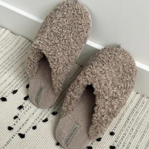 Peut inclure: Une paire de pantoufles beiges et duveteuses avec un motif boucl&eacute; textur&eacute;. Les pantoufles reposent sur un tapis &agrave; motifs avec des accents noirs et blancs. Les pantoufles portent l'inscription "Twins Home Style" sur le c&ocirc;t&eacute;.