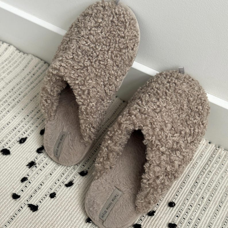 Peut inclure: Une paire de pantoufles beiges et duveteuses avec un motif boucl&eacute; textur&eacute;. Les pantoufles reposent sur un tapis &agrave; motifs avec des accents noirs et blancs. Les pantoufles portent l'inscription "Twins Home Style" sur le c&ocirc;t&eacute;.