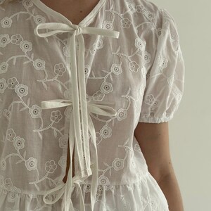 May include: White embroidered blouse with a floral pattern. The blouse features short, puffed sleeves and a tie-front closure. The delicate embroidery and lightweight fabric create a feminine and airy aesthetic, perfect for warm weather. The blouse has a ruffled hem.