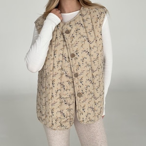 Può includere: Un gilet trapuntato beige con un motivo floreale blu navy, marrone e giallo. Il gilet ha una chiusura con bottoni e uno scollo tondo. Il gilet è sovrapposto a una maglia bianca a maniche lunghe.