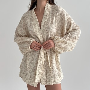 Op de afbeelding: Crèmekleurige kimono-achtige badjas met bloemenprint, lange mouwen en een striksluiting. De badjas heeft een delicaat patroon van kleine bruine bloemmotieven. De stof lijkt zacht en licht, geschikt om te loungen of te layeren.