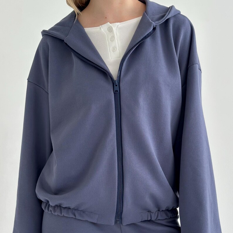 May include: A blue zip-up hoodie with a drawstring waist and a hood. The hoodie has a full-length zipper and long sleeves. A white Henley shirt is visible underneath. The hoodie is made of a soft material.