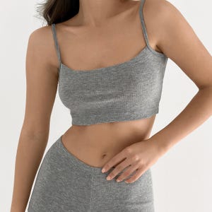 Op de afbeelding: Een grijze geribbelde crop top met een ronde halslijn en dunne spaghettibandjes. Het kledingstuk is gemaakt van een zachte, getextureerde gebreide stof die boven de taille valt en een nauwsluitend silhouet biedt met verticale ribbels.