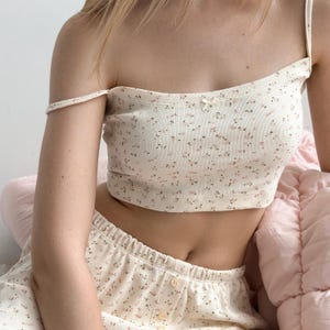 Op de afbeelding: Cr&egrave;mekleurige tweedelige pyjamaset met een delicaat bloemmotief in roze en bruin. De set bevat een crop top met dunne bandjes en een kleine strik, en bijpassende shorts. De stof lijkt geribbeld.