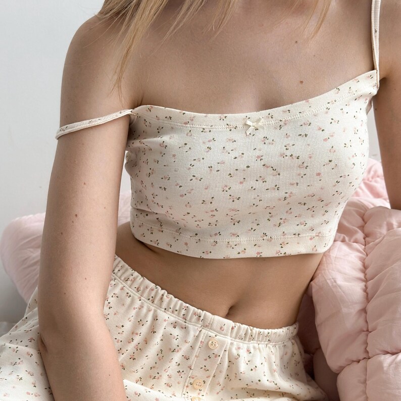 Op de afbeelding: Cr&egrave;mekleurige tweedelige pyjamaset met een delicaat bloemmotief in roze en bruin. De set bevat een crop top met dunne bandjes en een kleine strik, en bijpassende shorts. De stof lijkt geribbeld.