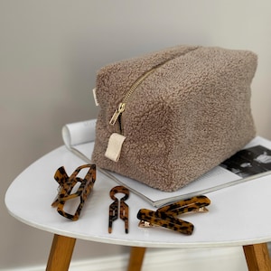 Puede incluir: Un neceser beige y esponjoso con cremallera dorada y varias pinzas para el pelo de carey. La bolsa es rectangular y está sobre una mesa blanca junto a una revista. Las pinzas son de varios tamaños.