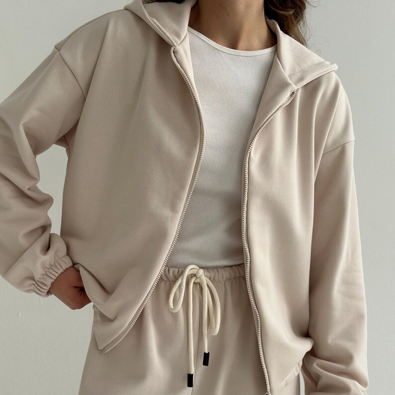 May include: A beige zip-up hoodie with a matching drawstring sweatpants. The hoodie has a hood and ribbed cuffs. A white ribbed tank top is visible underneath the jacket. The drawstring on the sweatpants has black tips. The outfit is made of a soft material.