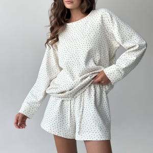 May include: Cream-colored loungewear set featuring a long-sleeved top and matching shorts. The set is adorned with a delicate, all-over floral pattern in a muted green hue. The shorts have a drawstring waist. The fabric appears soft and comfortable, perfect for relaxing.