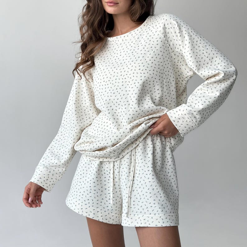 May include: Cream-colored loungewear set featuring a long-sleeved top and matching shorts. The set is adorned with a delicate, all-over floral pattern in a muted green hue. The shorts have a drawstring waist. The fabric appears soft and comfortable, perfect for relaxing.
