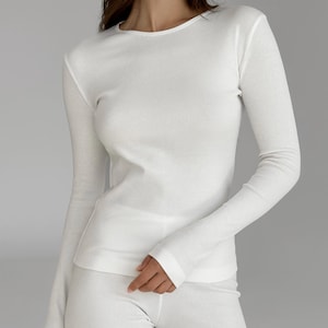 Könnte beinhalten: Ein weißes Langarmshirt mit feiner Rippstruktur, einem schlichten Rundhalsausschnitt und einer körpernahen Passform. Das Kleidungsstück besteht aus einem leichten Material mit einem durchgehenden vertikalen Rippmuster an Ärmeln und Rumpf.