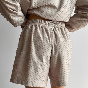 Puede incluir: Conjunto de dos piezas beige con una parte superior de manga larga y pantalones cortos a juego. La tela tiene un patr&oacute;n texturizado con puntos. Los pantalones cortos tienen cintura el&aacute;stica. El conjunto es de color neutro.