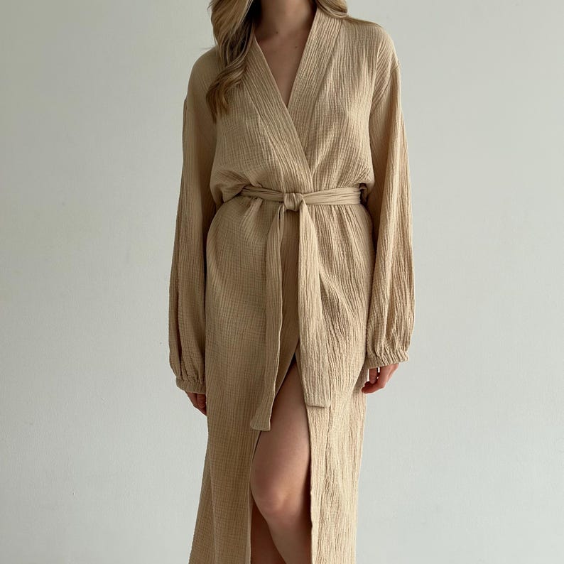 May include: A long, beige robe with a V-neck and long sleeves. The robe is tied at the waist with a matching fabric belt. The fabric has a textured appearance. The robe has a high slit on the left side.