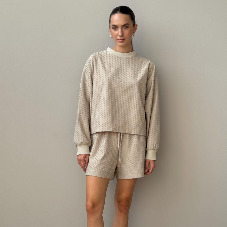 Puede incluir: Conjunto beige de top de manga larga y shorts texturizados. El top tiene cuello redondo y pu&ntilde;os acanalados. Los shorts tienen cintura con cord&oacute;n. La tela tiene un estampado de puntos. El conjunto es de color neutro.
