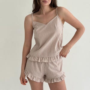 May include: A two-piece pyjama set featuring a sleeveless camisole top and matching shorts. Both pieces are made of a light beige fabric with thin white vertical stripes. The top has thin shoulder straps and a ruffled hem, while the shorts feature a ruffled hemline.