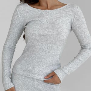 Op de afbeelding: Een lichtgrijs geribd shirt met lange mouwen, een ronde hals en drie kleine knopen. Het shirt heeft een getailleerd silhouet en is gemaakt van een zachte, getextureerde stof. De mouwen zijn lang en eindigen bij de polsen.