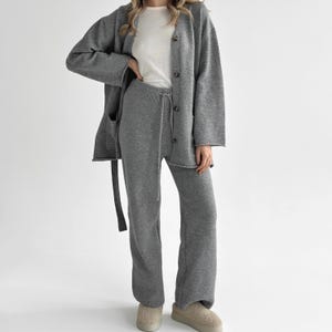 Op de afbeelding: Een grijs gebreid vest met knoopsluiting aan de voorzijde en een bijpassende gebreide broek met wijde pijpen en een trekkoord in de taille. De outfit is gecombineerd met een effen wit T-shirt en beige instapschoenen voor een gecoördineerde set.