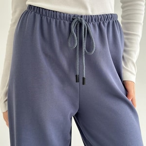 May include: Close-up of blue sweatpants with a drawstring waist. The pants are a solid color and appear to be made of a soft material. The drawstring is a lighter shade of blue with black aglets.