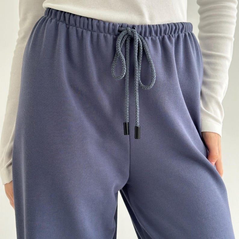 May include: Close-up of blue sweatpants with a drawstring waist. The pants are a solid color and appear to be made of a soft material. The drawstring is a lighter shade of blue with black aglets.