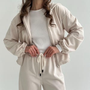 May include: A beige two-piece tracksuit featuring a zip-up hooded jacket and matching drawstring pants. The jacket has a white undershirt visible underneath. The outfit is made of a soft, comfortable material.